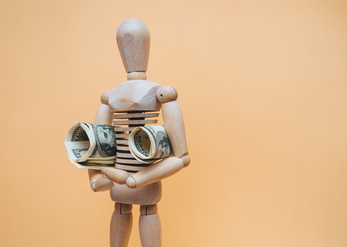 A fanny wooden mannequin holds money (dollars) under his arms. Theft concept, financial pyramid, lottery winning, money back, value added tax refund. Donation.