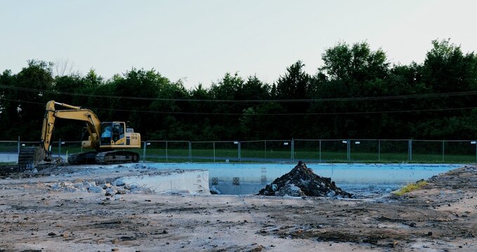 Yellow excavator tearing down abandoned public swimming pool, showing crushed concrete, rebar, rubble during urban redevelopment demolition dirty zone