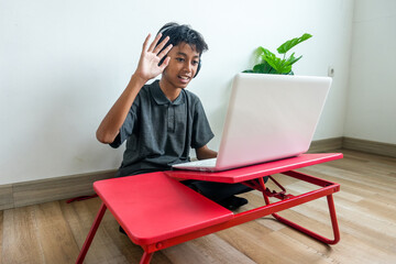 Asian Teen Boy Waving Hello During an Online Video Call on His Laptop at Home © Gatot