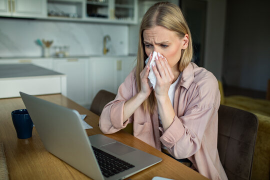 Sick woman sneezing into tissue while working on laptop at home