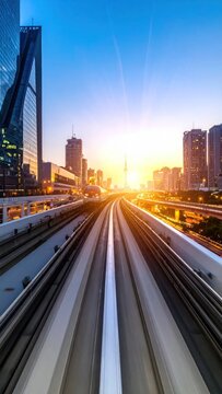 Elevated train tracks lead toward a bright sunset in a bustling city with tall skyscrapers