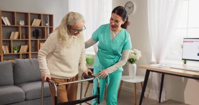 Nurse supports senior woman with walker. Caregiver guides the patient in homecare, providing support and improving mobility during rehabilitation in a living room. Kind home aid for seniors.