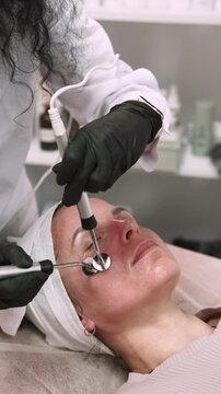Vertical video. Woman treatment. Microcurrent procedure. Professional cosmetologist in black gloves using device for facial skin lifting.