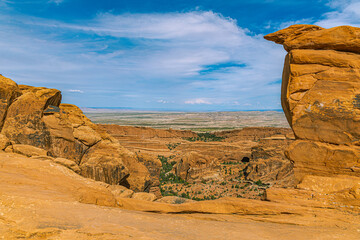A vast desert valley and distant mesas stretch below a rocky overlook framed by sandstone boulders in Arches National Park.  © ZL Visuals