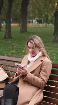 Vertical video. Phone communication. Resting outdoor. Female in beige fashion trench using online app on wooden bench in autumn park.