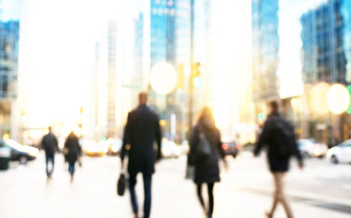 Obraz premium A blurred urban scene depicting people walking in a city with tall glass buildings and bright sunlight, conveying a busy metropolitan atmosphere.