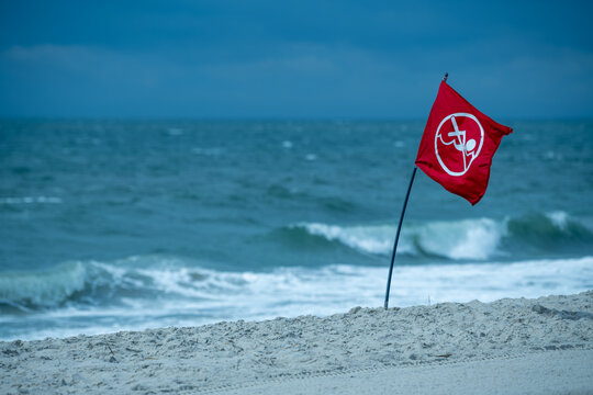 A red no-swimming warning flag with a white prohibition symbol flies on a pole planted in beach sand, with rough, churning surf and a moody blue-gray sky in the background at Cape May, New Jersey.