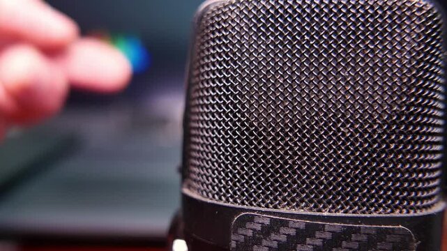 POV (zoom in): man speaking into a professional microphone while gesturing during a podcast episode recording. In the background, colorful sound waves move on a laptop screen. Voiceover recording.