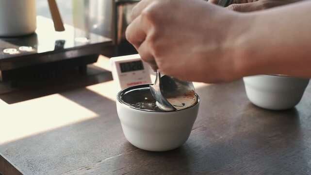 Barista using two spoons to skim the foam and coffee grounds from the surface of a cupping bowl during a sensory evaluation.