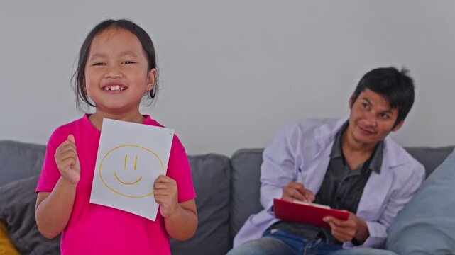 Cheerful child patient holding happy face card as male doctor writes on clipboard behind