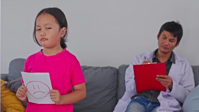 Sad child patient holding smiley card and turning away from male doctor writing on clipboard