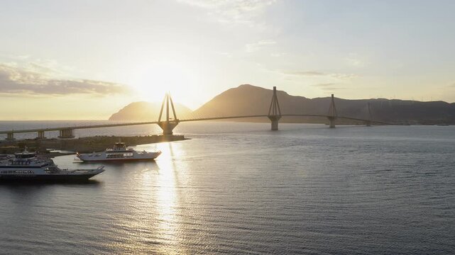 Rio-Antirrio Bridge over Gulf of Patras at sunset near Patra Greece, drone dolly backlit glow above yacht ferries, mountain ridgeline