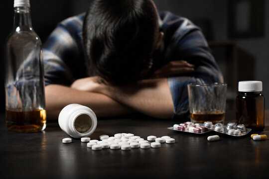 A person is slumped over a dark surface with pills and bottles scattered around them.