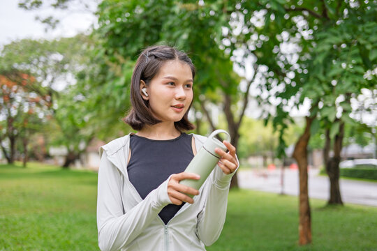 Asian Gen Z woman using reusable stainless tumbler after jogging in green urban park for sustainable wellness lifestyle