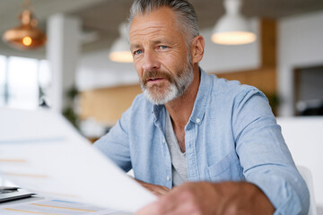 Fototapeta premium Mature man studies documents at a modern desk. His focused gaze suggests deep concentration. Soft lighting highlights calm, thoughtful expression. Professional setting implies business or planning