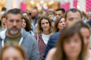 Fototapeta premium Woman stands out in crowded shopping mall crowd. She gazes calmly, surrounded by blurred shoppers. Lighting highlights her from warm overhead fixtures. Scene captures urban life's quiet intensity