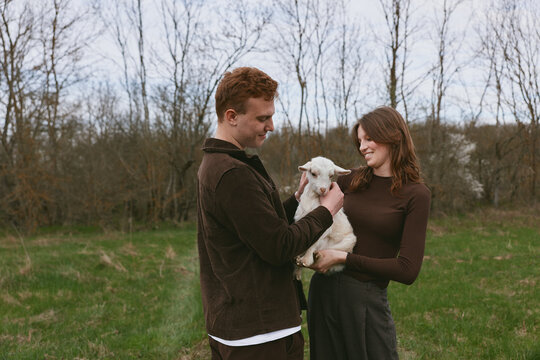 A couple outdoors in a field interact with a goat kid, showing affection, smiles, and gentle care, capturing a warm lifestyle moment with animals and natural scenery in daylight and companionship