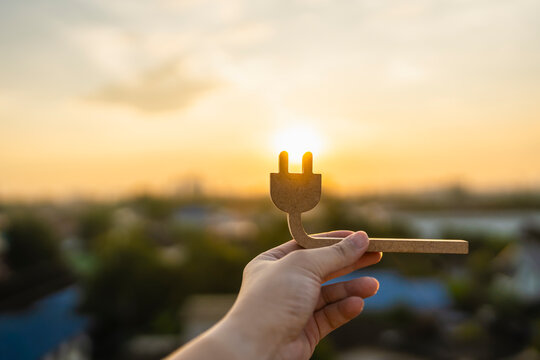 Green energy saving concept with hand holding wooden electric plug silhouette against sunset sky for renewable power, sustainable eco ecosystem and technology.