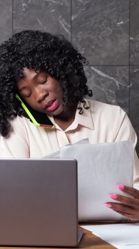 Young black businesswoman simultaneously engaging in a phone conversation, working on her laptop, and organizing financial paperwork, showcasing a busy and efficient multitasking work style