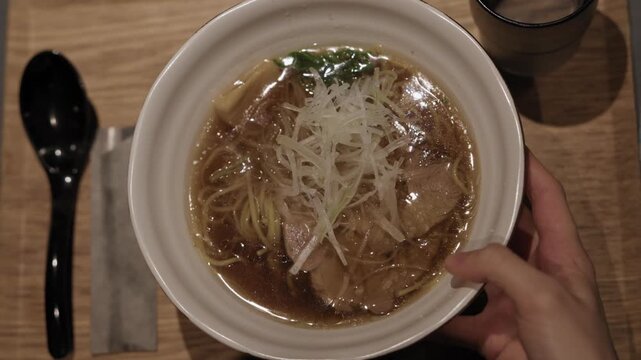 Hand slides bowl of shoyu ramen with chashu pork and leek onto wooden table