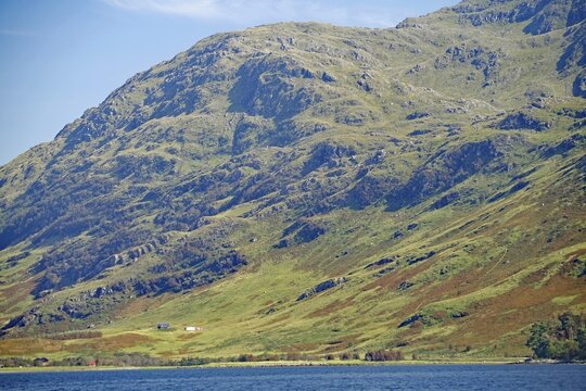 Green slopes of a large mountain with scattered farm buildings under a clear sky, Knoydart, Highlands, Scotland, United Kingdom