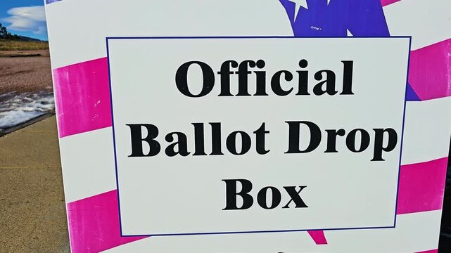 Close view of an official ballot drop box wrapped in American flag artwork, positioned beside a public sidewalk with snow patches for secure absentee and mail-in US election voting outdoors.