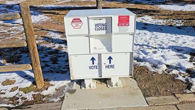 Outdoor mail-in ballot drop box marked Vote Here and Voting Closed, with secure election collection slots, warning signs, directional arrows, snow, and winter sunlight at a polling location.