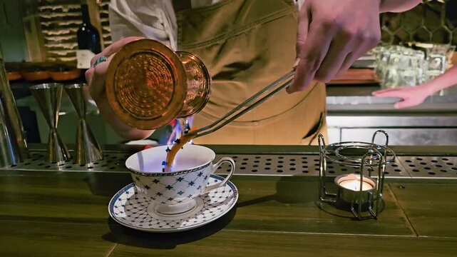 Bartender pours flaming liquid from a copper pot into a patterned teacup, with blue flame, candle warmer, metal tongs, warm bar lighting, and dramatic tea cocktail service.