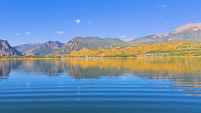 Scenic Dillon, Colorado, lake view with a panning camera, vivid yellow aspens, evergreen hillsides, rocky peaks, shoreline buildings, blue sky, and bright autumn sunlight.