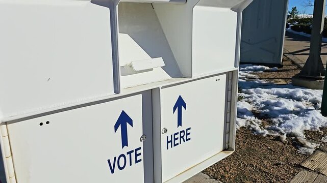 White ballot drop-off box for mail-in election voting displays Vote Here arrows, secure collection compartments, warning notices, and a Voting Closed sign at a snowy outdoor polling site.