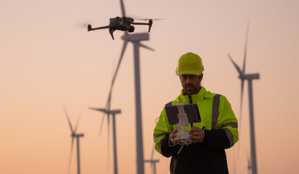 Engineers male flying drone surveying and checking wind turbines from the high angle view of the field during beautiful sunset. using drone technology for work.