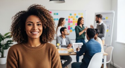 Fototapeta premium Portrait of a confident young woman with curly hair smiling in a modern office with colleagues collaborating in the background