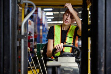 A tired female forklift operator sitting on her vehicle, touching her forehead with her hand. She is wearing a safety vest and black shirt © offsuperphoto