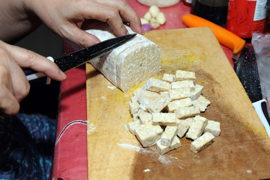 person slicing fresh raw tempeh into small rectangular cubes using a black knife on a wooden cutting board.