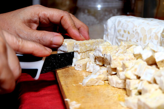 side view, person slicing fresh raw tempeh into small rectangular cubes using a black knife on a wooden cutting board.