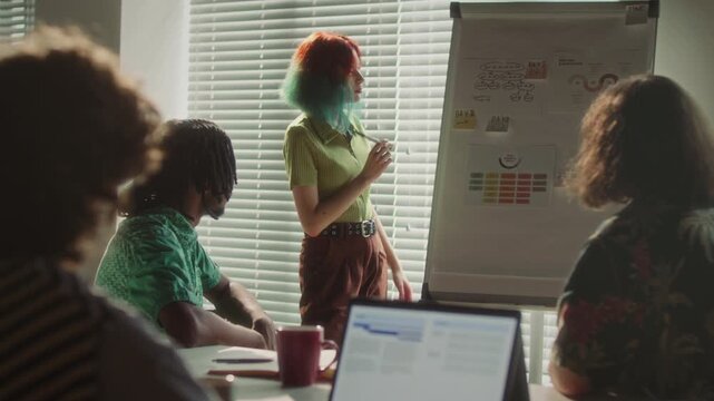 Young woman with colorful hair explaining printed infographics on flip chart to diverse team of colleagues while leaning business presentation during office meeting
