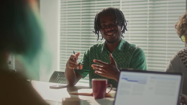 Young smiling Black businessman explaining his ideas to diverse coworkers during team meeting in sunlit office. Over the shoulder view
