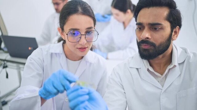 Female Scientists in eyeglasses and male scientists looking closely at green sample held in gloved hands, focused scientific examination in busy laboratory.