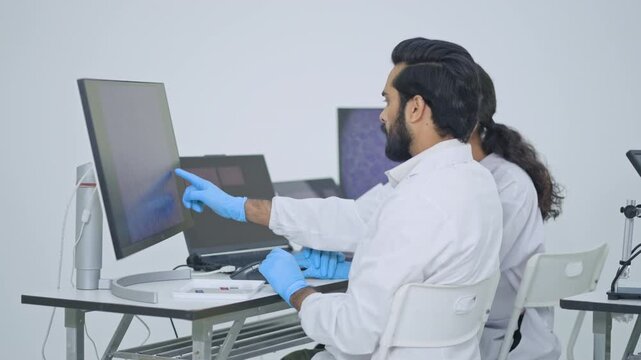 Researcher in gloves working at desk with monitors showing microscopy scans, colleague besidpharmaceuticale him, digital analysis in lab.