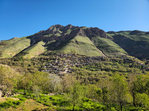 A terraced mountain village nestles among dense green orchards, framed by rugged rocky peaks rising sharply against a clear blue sky.
📍 Kamalla village, Hawraman Takht, Kurdistan 