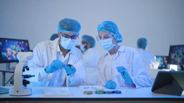 Male and female scientists in masks and goggles inspecting small sample tube at bench with petri dishes and tablet, lab research in blue light.