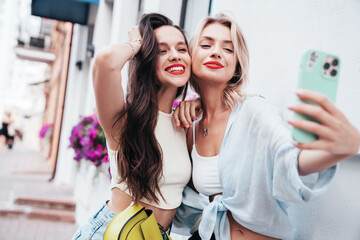 Two young beautiful smiling female in trendy summer clothes. Sexy carefree women posing in the street. Positive models having fun, hugging in sunny day. With bright makeup and red lips, take selfie © halayalex
