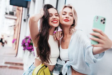Two young beautiful smiling female in trendy summer clothes. Sexy carefree women posing in the street. Positive models having fun, hugging in sunny day. With bright makeup and red lips, take selfie © halayalex