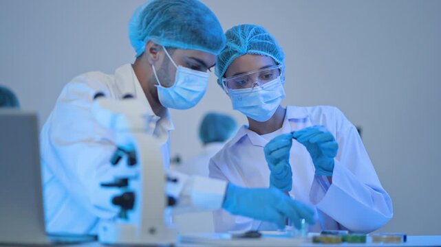 Two researchers in protective caps and masks examining tiny specimen together, microscope in foreground, focused laboratory work.