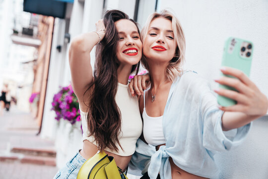 Two young beautiful smiling female in trendy summer clothes. Sexy carefree women posing in the street. Positive models having fun, hugging in sunny day. With bright makeup and red lips, take selfie