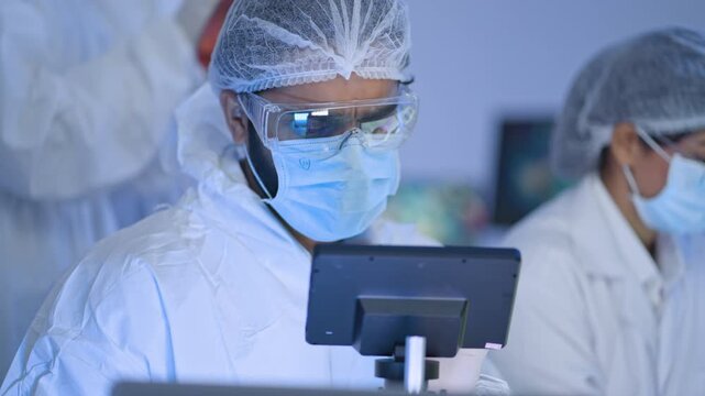 Specialist in white coverall, mask and reflective goggles operating touchscreen device, colleagues working in cleanroom production facility.
