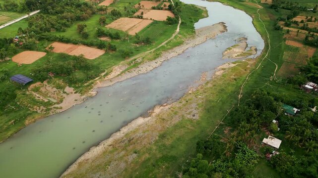 Aerial flyover of a shallow river amid surrounding farmlands, grassy banks, and lush tropical greenery in Pajo, Virac, Catanduanes, Philippines
