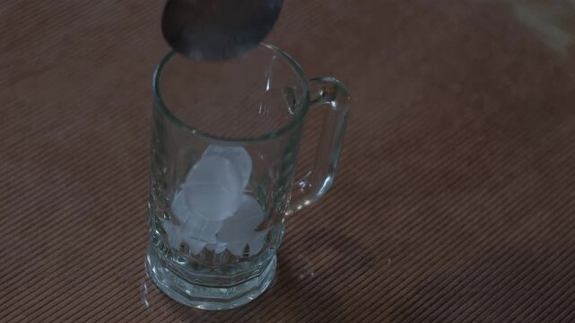 Medium static shot of ice balls being dropped into a clear glass mug on a tabletop for cold coffee or drink preparation indoors