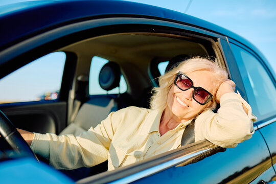 Happy senior woman wearing sunglasses and smiling, enjoying retirement freedom while driving a car on a beautiful day