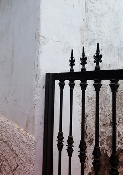A classic black wrought iron fence with ornate fleur-de-lis finials stands against a weathered white plaster wall. 